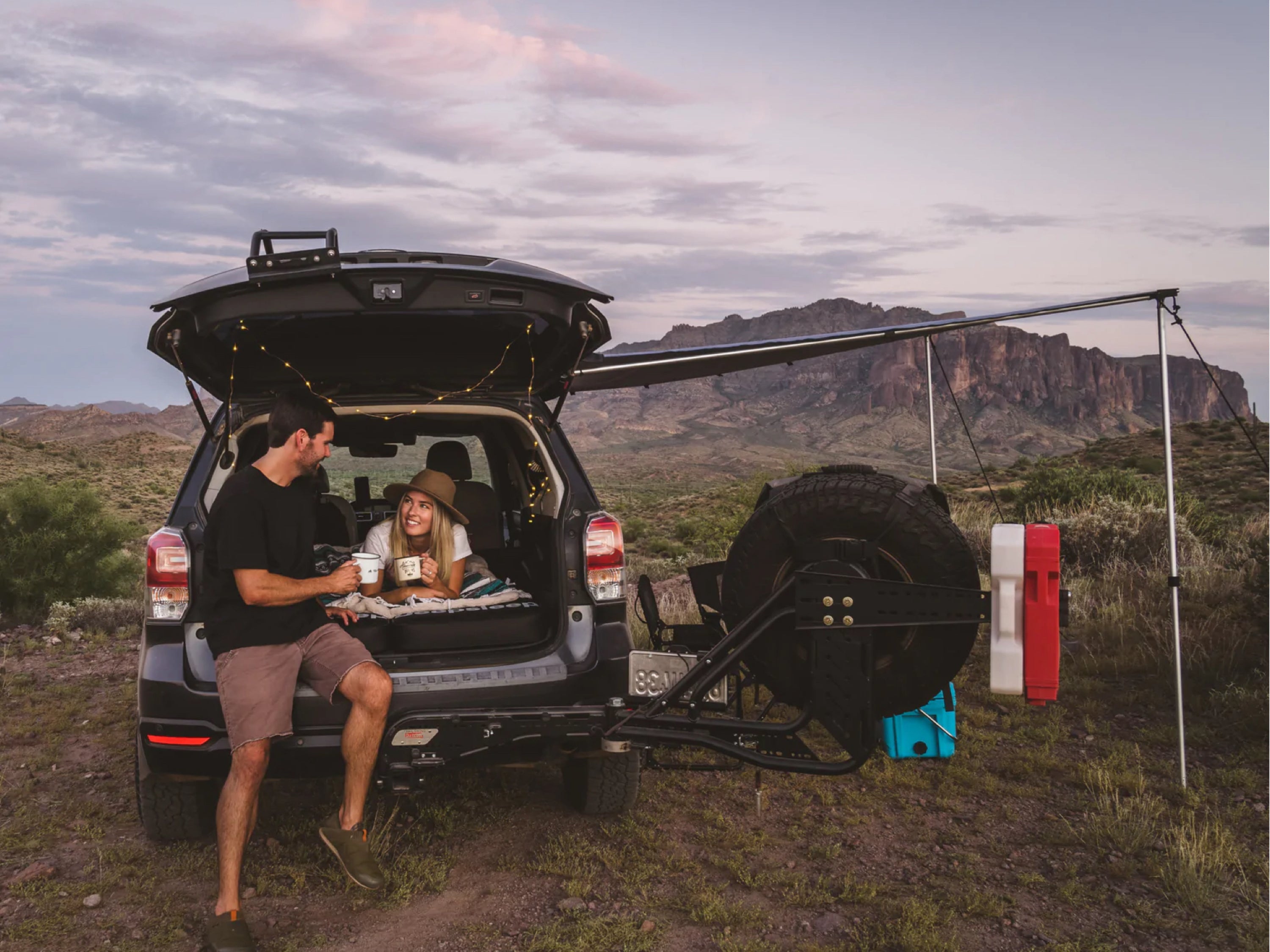 Male and female holding mugs while car camping on foam mattresses in the back of overland SUV in the mountainous desert at dusk