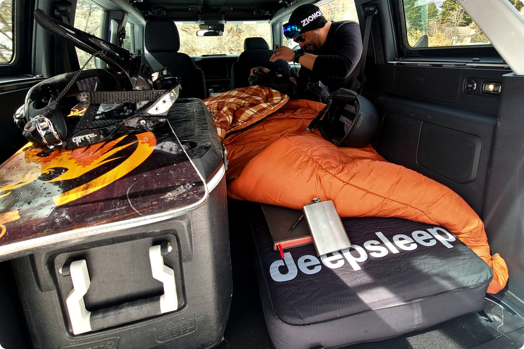 Closeup inside interior overland vehicle outfitted for car camping with Deepsleep solo foam mattress, sleeping bag, cooler, skiing accessories and rugged man near passenger side door.