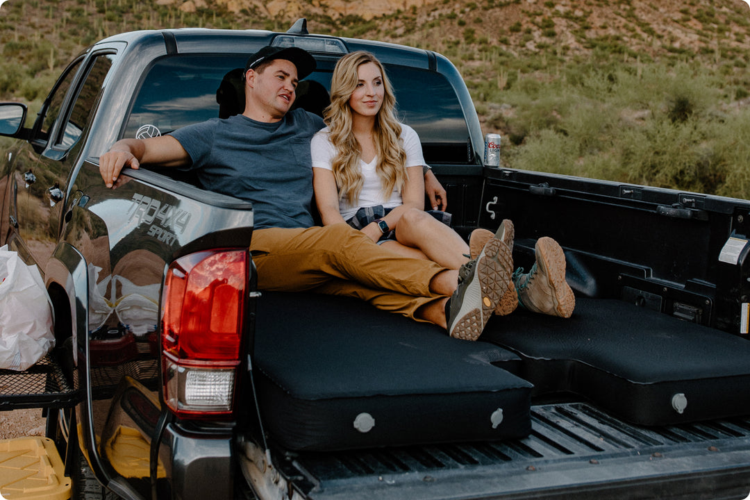 Male and blonde female sitting on an air mattress in the bed of a black Toyota Tacoma parked in the Arizona desert